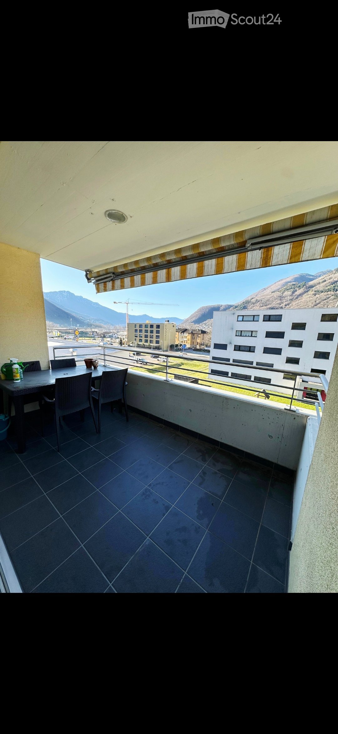 Balcony with tiled floor, metal railing, table, chairs, and a view of mountains and town.