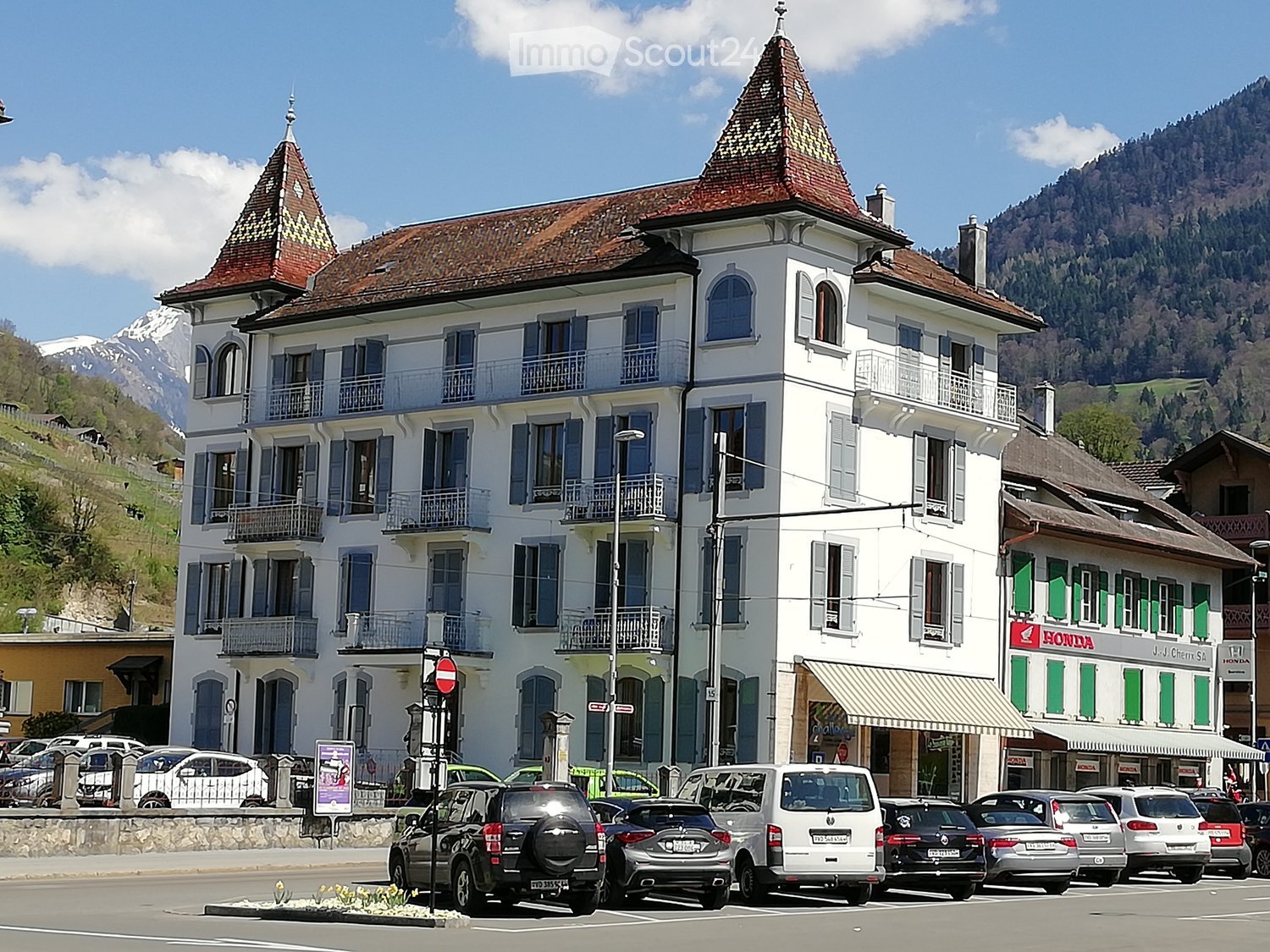 A large, white, multi-story building with blue shutters and balconies, featuring a distinctive red-tiled roof and ornate architectural details. The building is situated in a scenic area with mountains in the background and several parked cars in front.