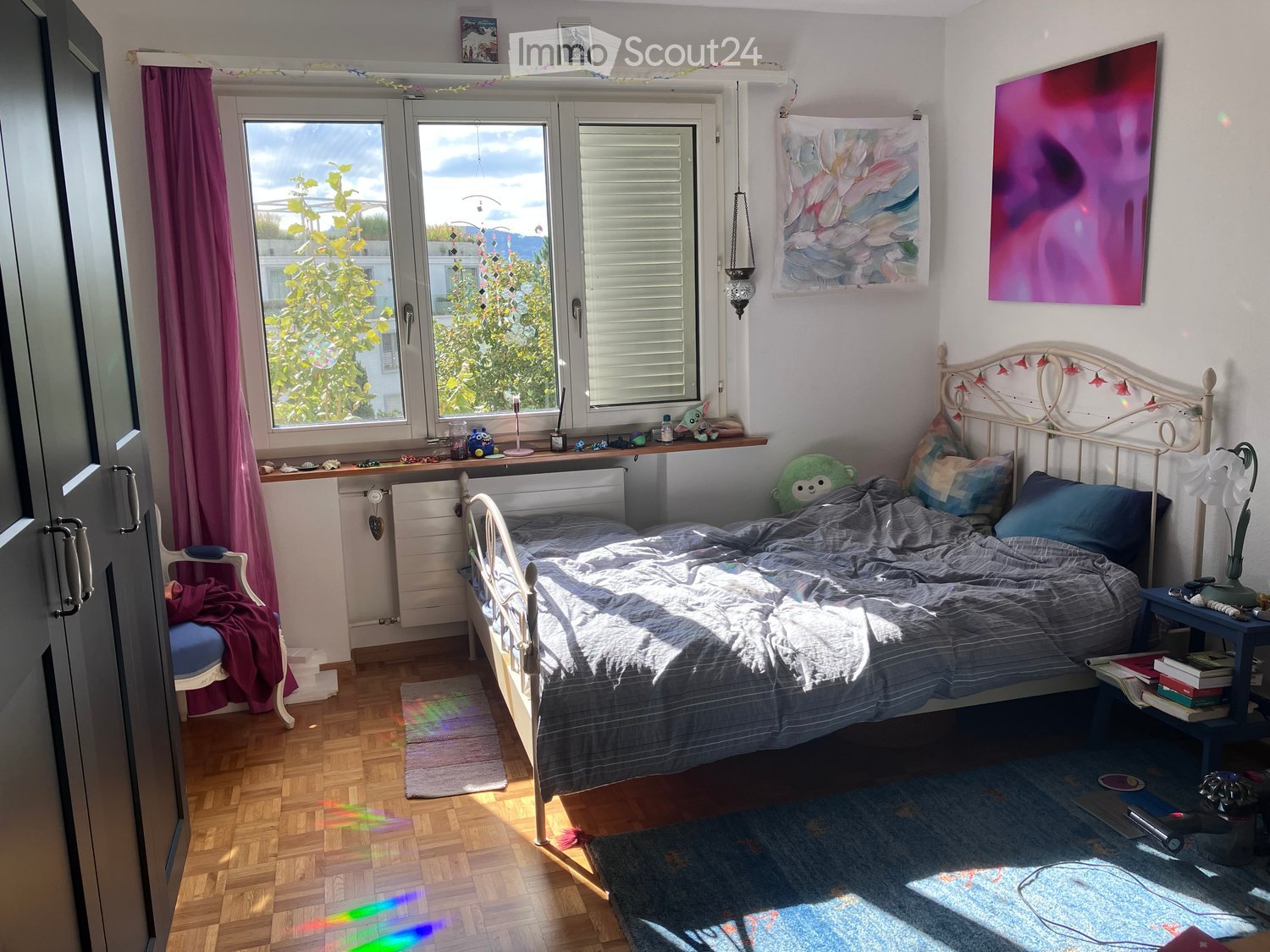 Cozy bedroom with wooden flooring, a white metal bed with a decorative headboard, a nightstand with books and a lamp, a small chair with a cushion, and a colorful painting on the wall. Natural light streams in through large windows with white blinds and p