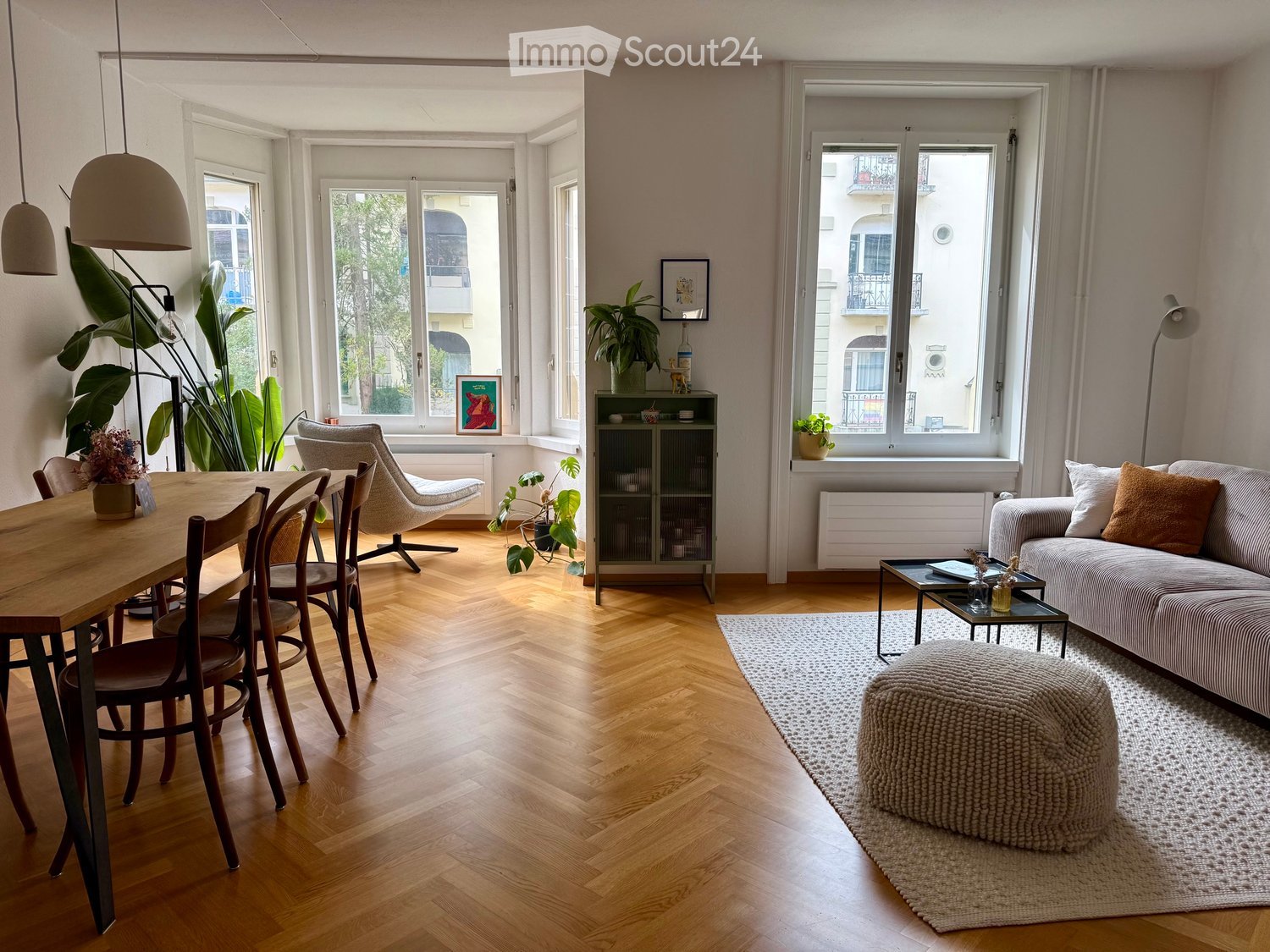 Bright living and dining area with wooden flooring, large windows, a sofa, ottoman, coffee table, dining table with chairs, and decorative plants.