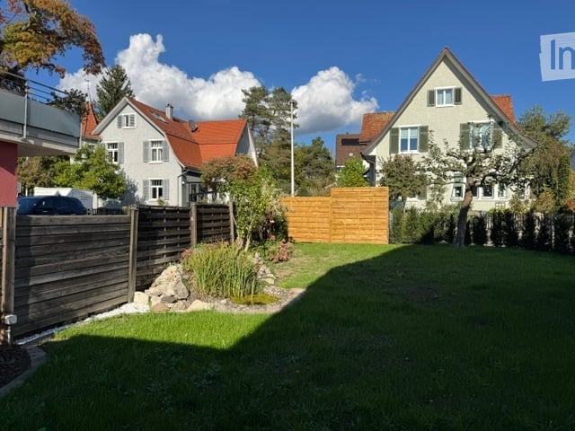 Well-maintained garden with a wooden fence, neatly trimmed grass, small shrubs, and a few trees. The background shows neighboring houses with red-tiled roofs and white walls.