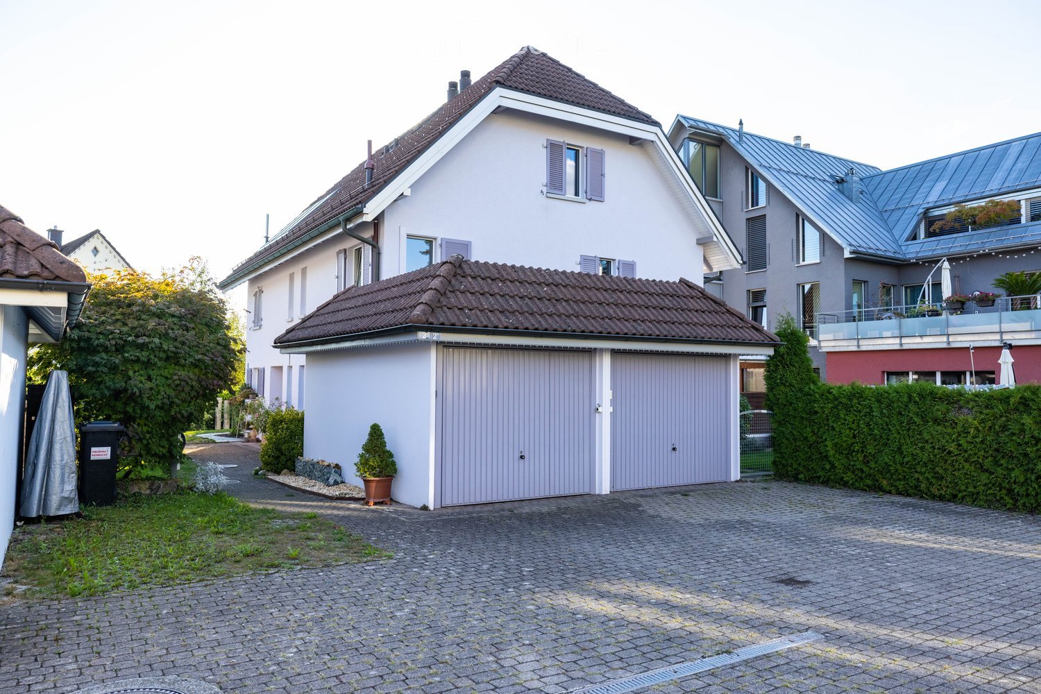 A two-story house with a garage, surrounded by greenery and a paved driveway.