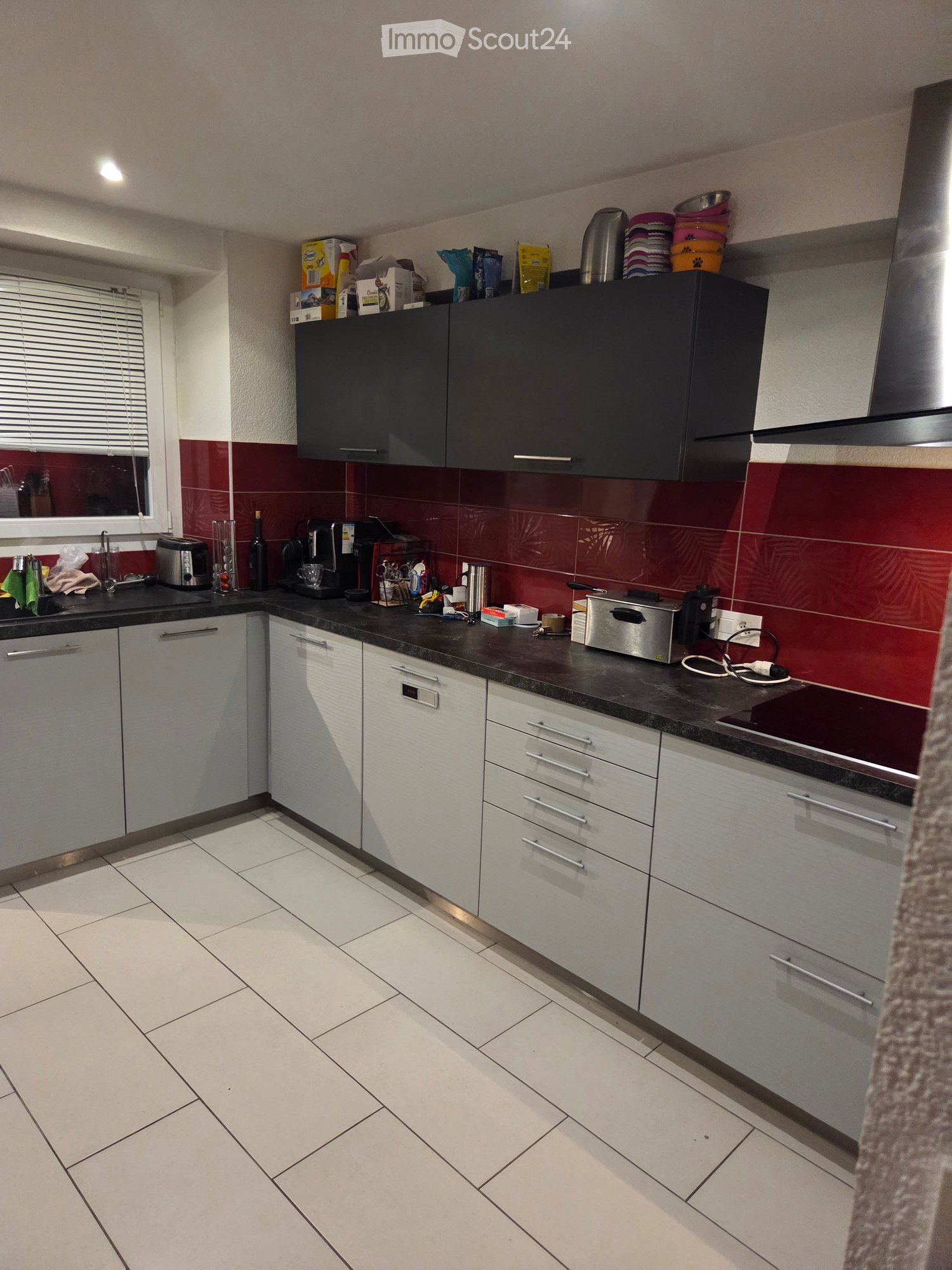 Modern kitchen with white tiled floor, white and black cabinets, black countertop, and various appliances including a coffee maker, toaster, and oven. The backsplash is red with a leaf pattern. There is a window with blinds on the left side.