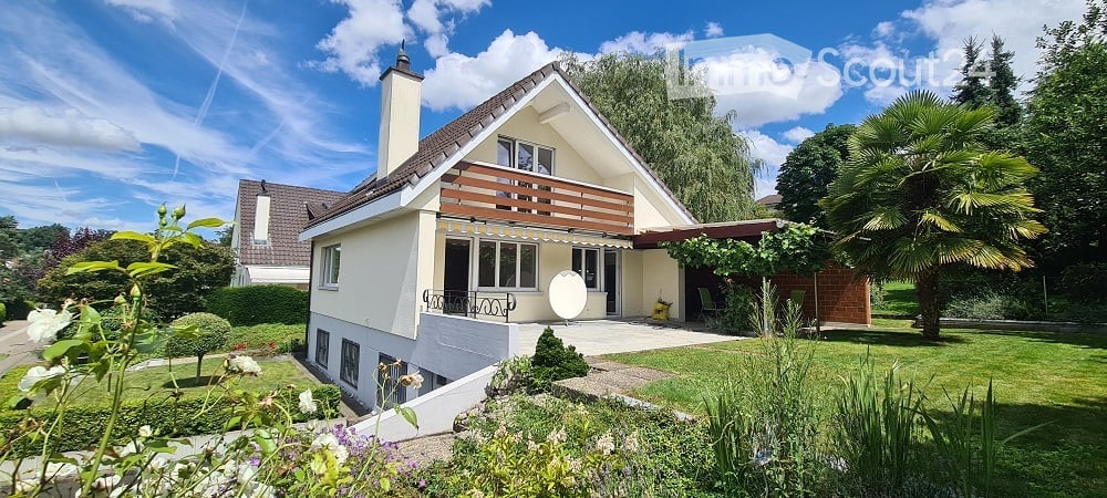 Two-story house with a modern design, featuring a balcony, large windows, and a well-maintained garden with various plants and trees.