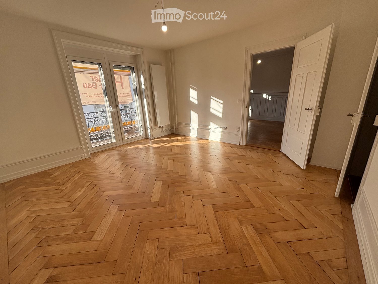 Empty room with wooden herringbone flooring, white walls, and large glass doors leading to a balcony.