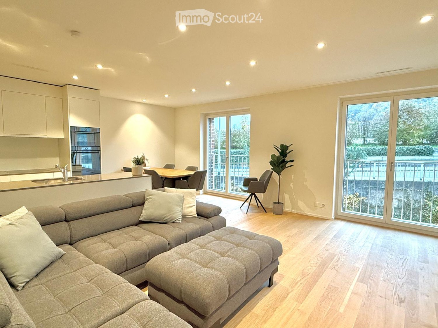 Open-plan living and dining area with a grey sectional sofa, coffee table, dining table with chairs, and a modern kitchen area with an oven and sink.