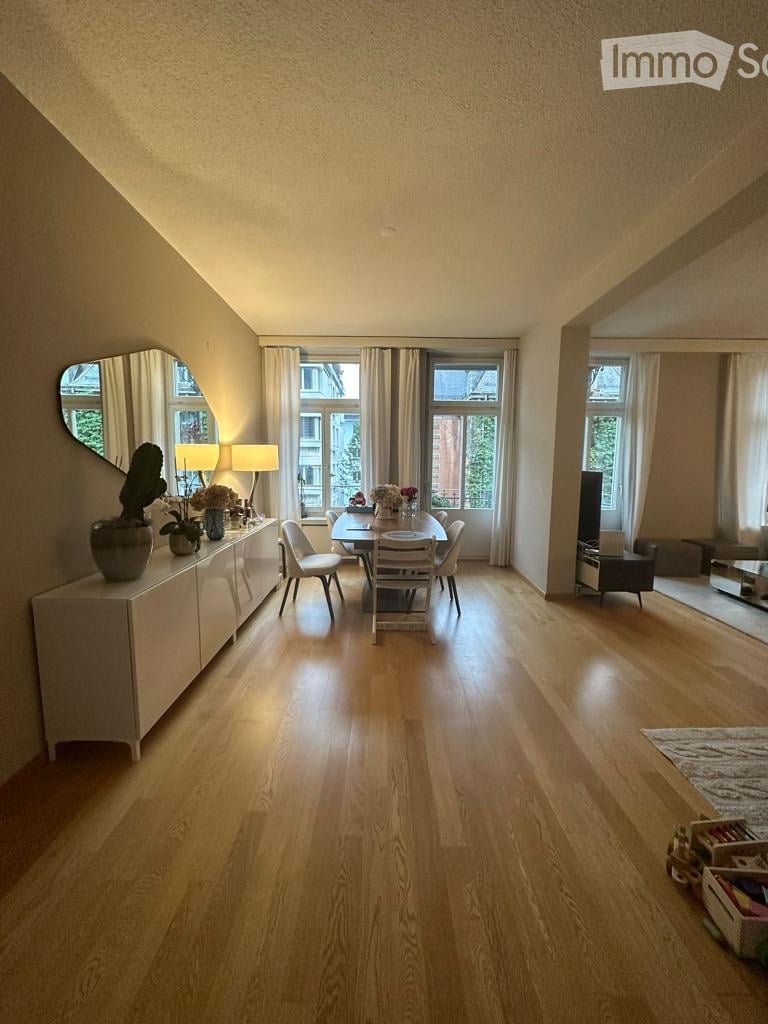Bright, open-plan dining and living area with wooden flooring, large windows, a dining table with chairs, a sideboard with decorative items, and a television in the background.