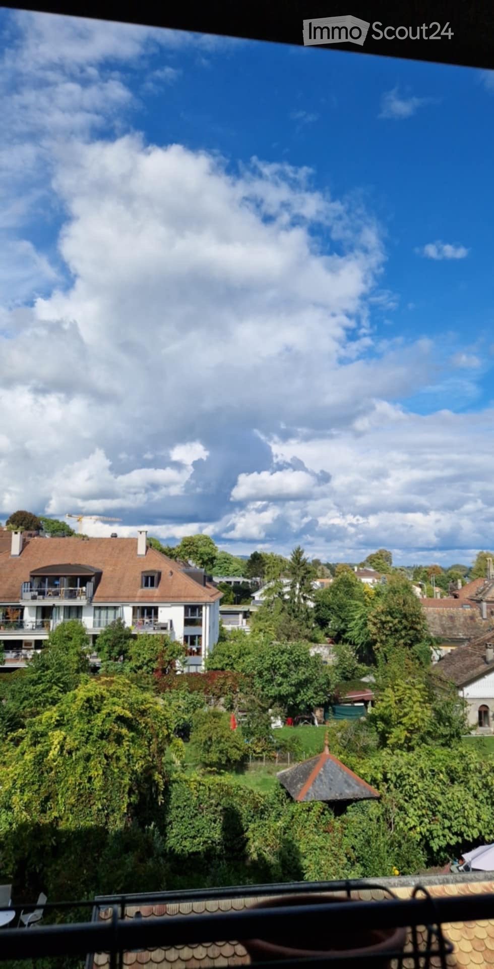 View from a balcony showing a residential area with houses, trees, and a clear blue sky.