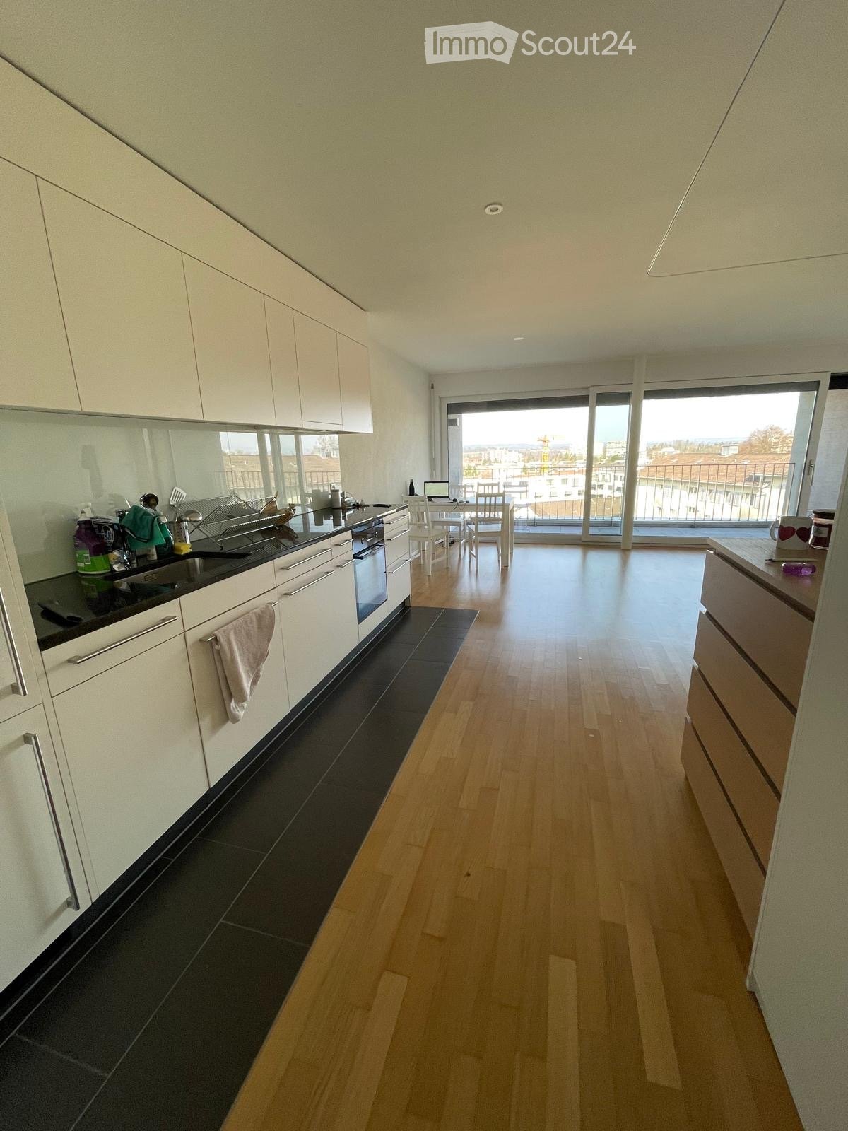 Modern kitchen with white cabinets, black countertop, and wooden flooring. Features an oven, sink, dish rack, and a small dining area with a table and chairs.