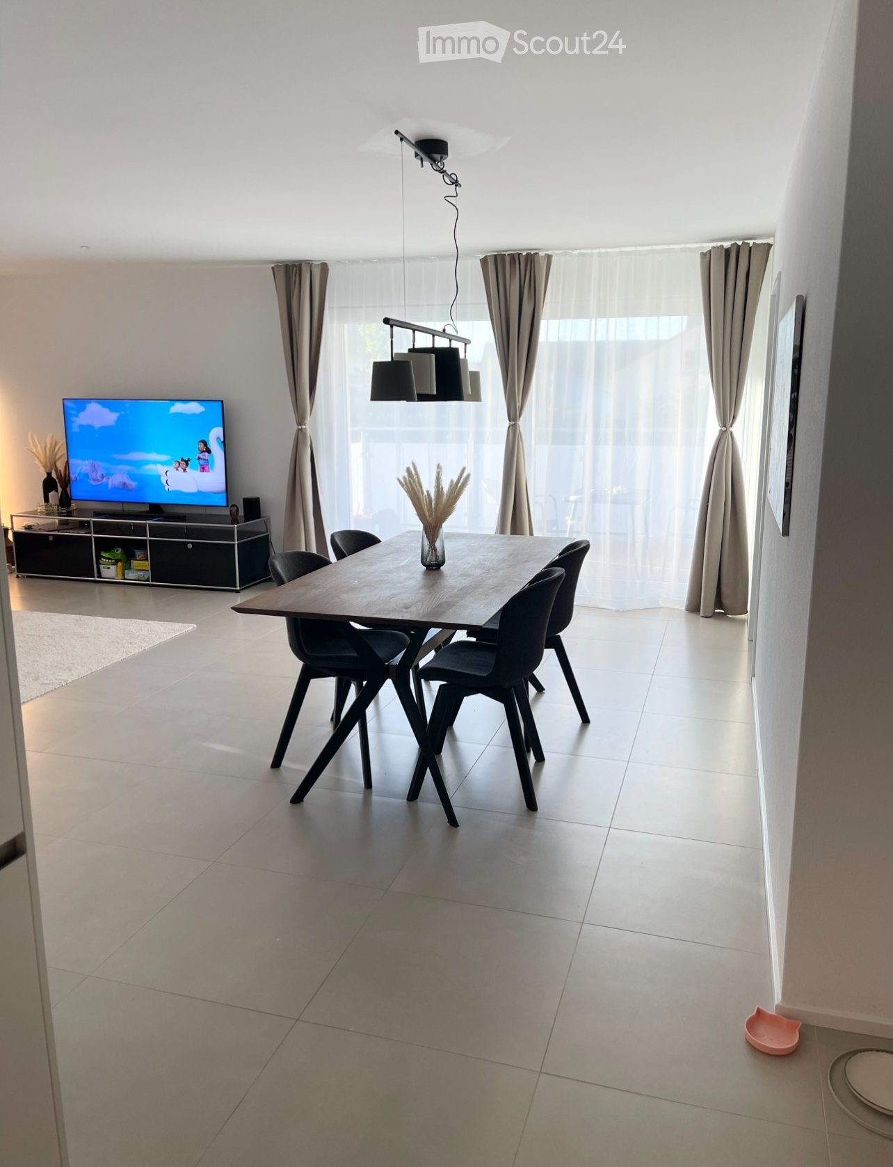 Modern dining area with a wooden dining table, black chairs, pendant lights, and a television in the background.