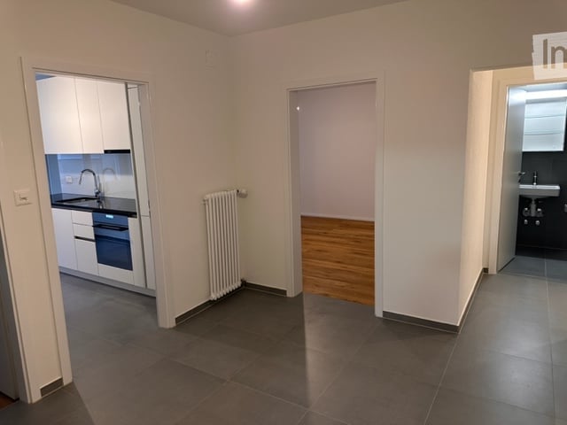 Empty room with wooden flooring, white walls, and a radiator. Adjacent rooms include a kitchen and a bathroom.