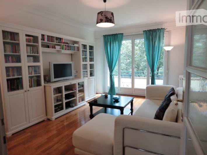 Bright living room with large windows and a view, featuring a white sectional sofa, a black coffee table, a television on a white cabinet, built-in bookshelves, and a floor lamp.