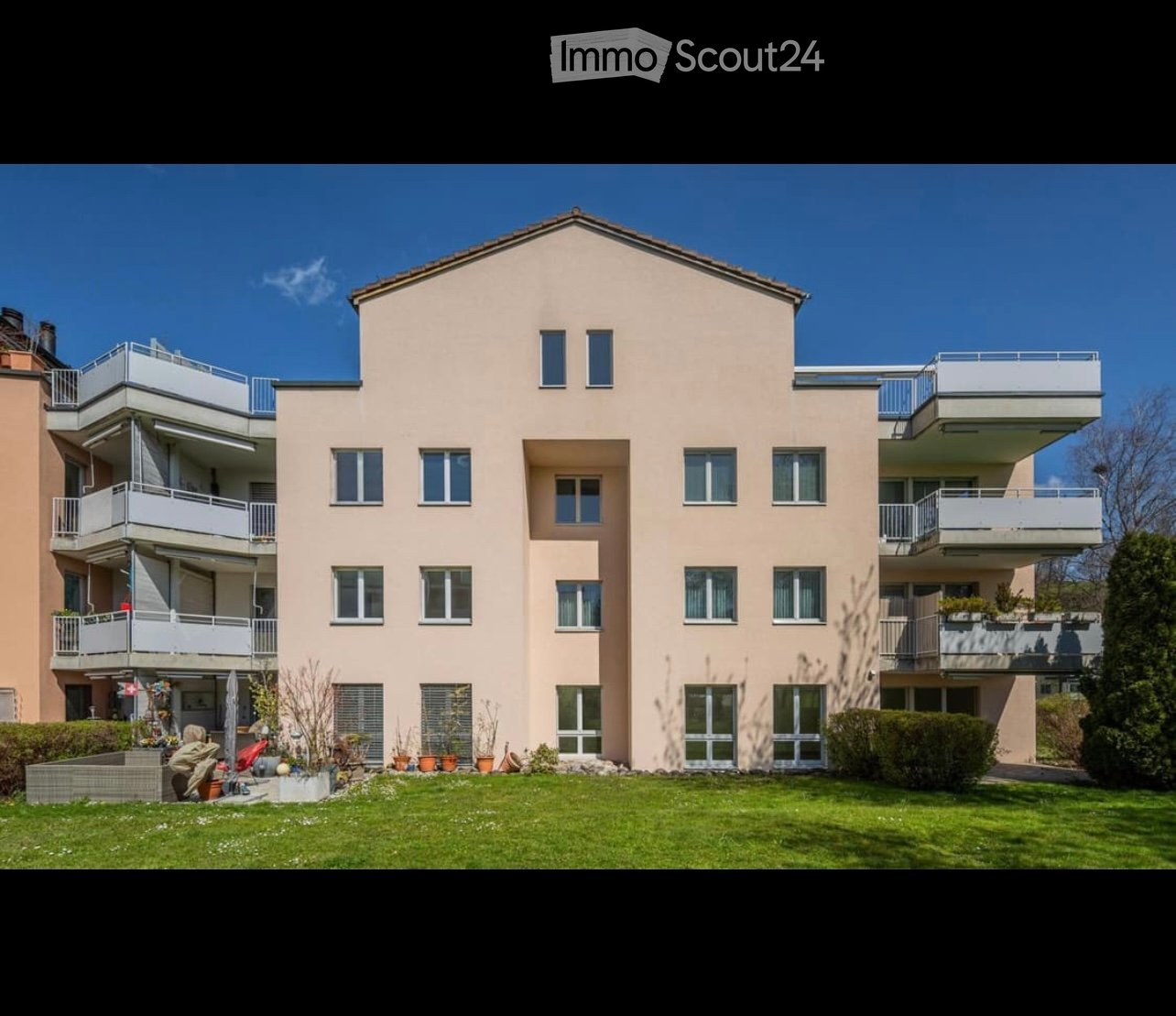 A modern, multi-story residential building with light beige walls, multiple windows, and balconies. The building is surrounded by a well-maintained lawn and small garden area with potted plants.