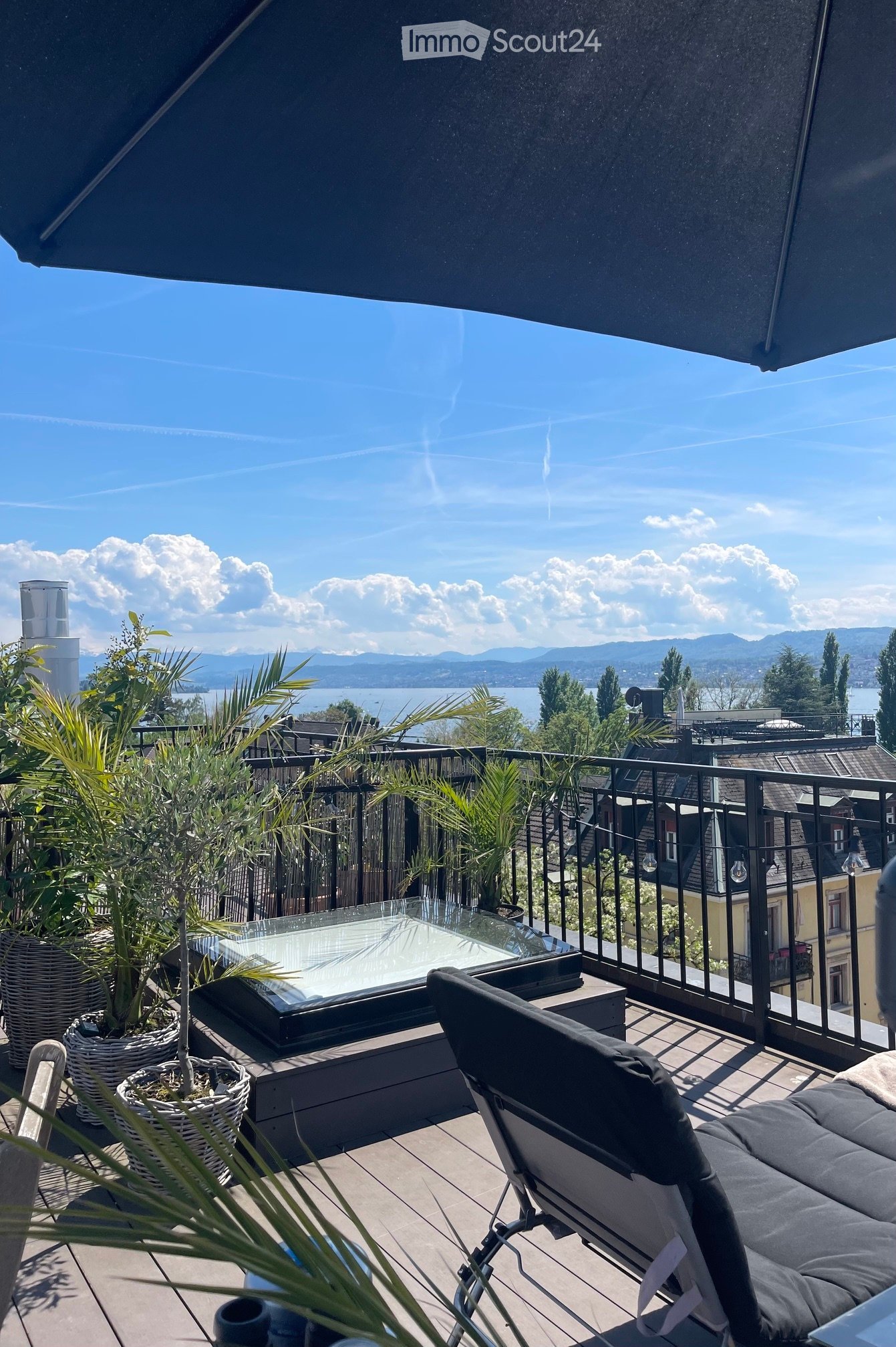 Balcony with a view of a lake and mountains, featuring a glass table, chairs, and potted plants.