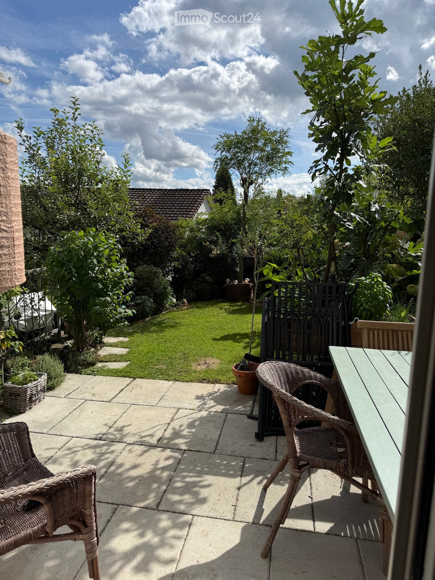 A well-maintained garden with a paved patio area featuring wicker chairs and a table, surrounded by lush greenery and trees.