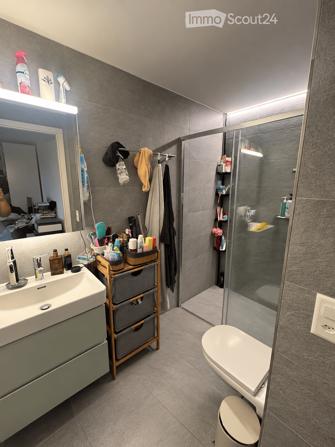 Modern bathroom with grey tiled walls and floor, featuring a white sink with a mirror, a toilet, a glass-enclosed shower, and a wooden storage unit with multiple drawers. Various toiletries and cosmetics are neatly arranged on the sink counter and in the
