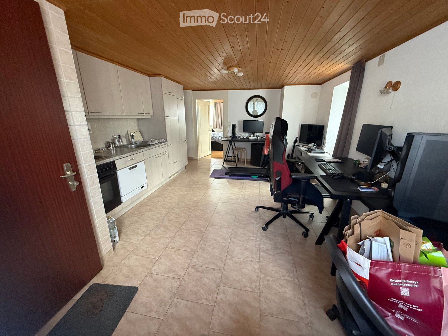 A furnished room with a kitchen area, a desk with a computer, and a chair. The kitchen features cabinets, a sink, and an oven. The room has a wooden ceiling and tiled floor.