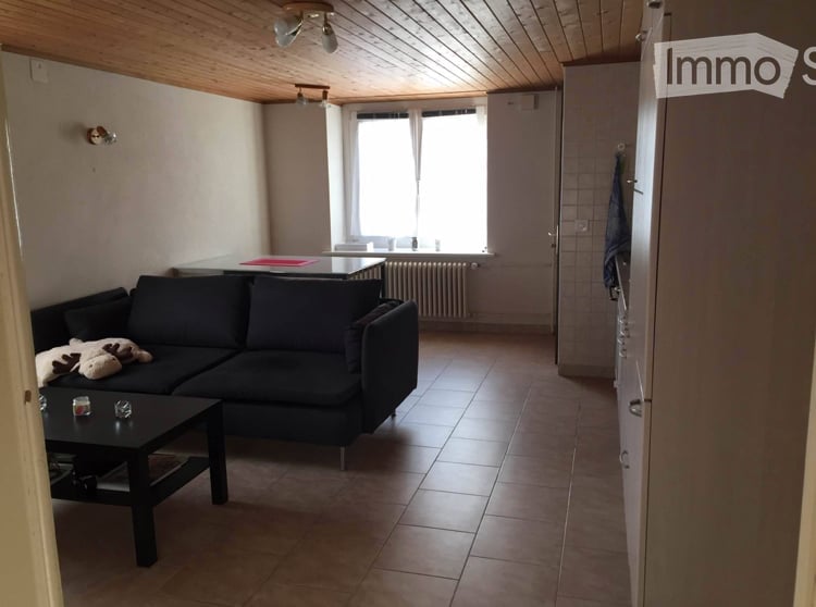 Living room with a black sectional sofa, a black coffee table, a dining table, and a radiator under the window. The room has wooden ceiling beams and tiled flooring.