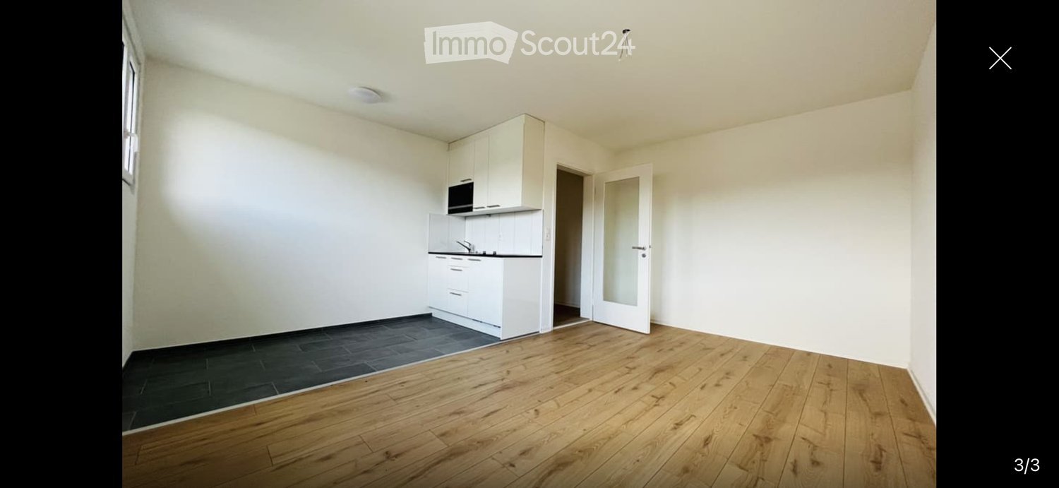 Empty room with wooden flooring, white walls, and a small white kitchen area with cabinets and a microwave. The room has a large window and a door leading to another space.