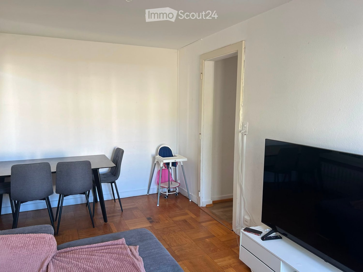 A modern living room with a grey sofa, a small dining table with four chairs, a baby chair, and a flat-screen TV on a white stand. The room features wooden flooring and white walls.