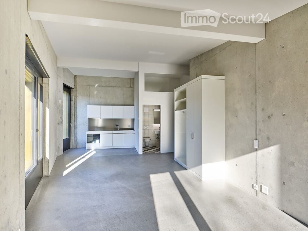 Modern, minimalist interior with white kitchen cabinets, a sink, a toilet, and a tall white cupboard. The space features concrete walls and floors with ample natural light from large windows.