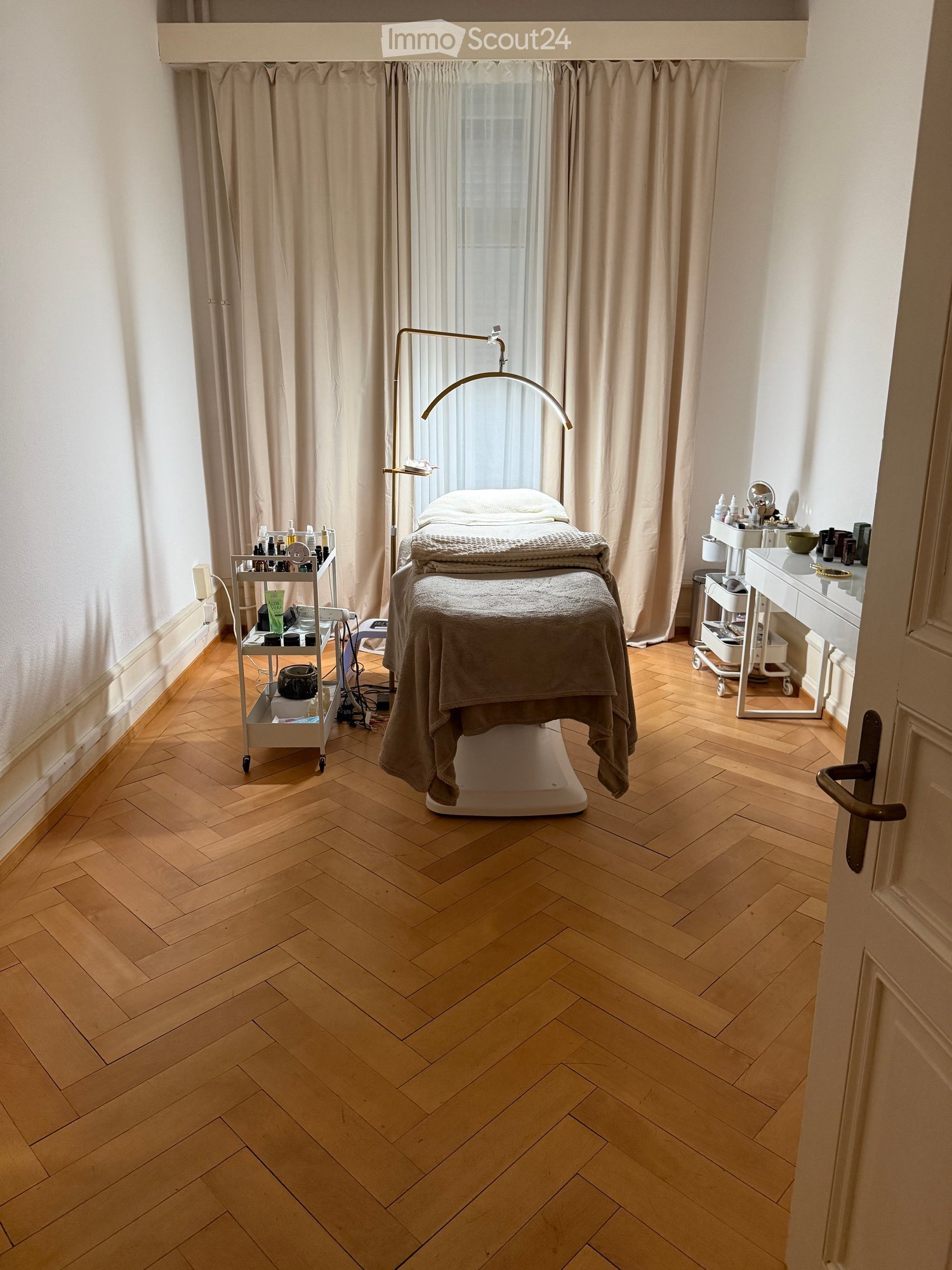 A well-lit, organized treatment room with a wooden floor, a treatment table covered with a blanket, a cart with various items, a cabinet, and a mirror.