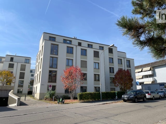 Modern multi-story apartment building with white facade, multiple windows, and surrounding greenery. Several cars are parked in front.