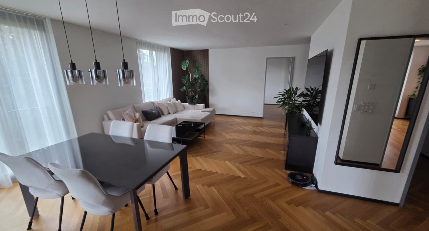 Modern living and dining area with light wooden flooring, a grey sectional sofa, a black coffee table, a glass dining table with white chairs, pendant lights, and plants.