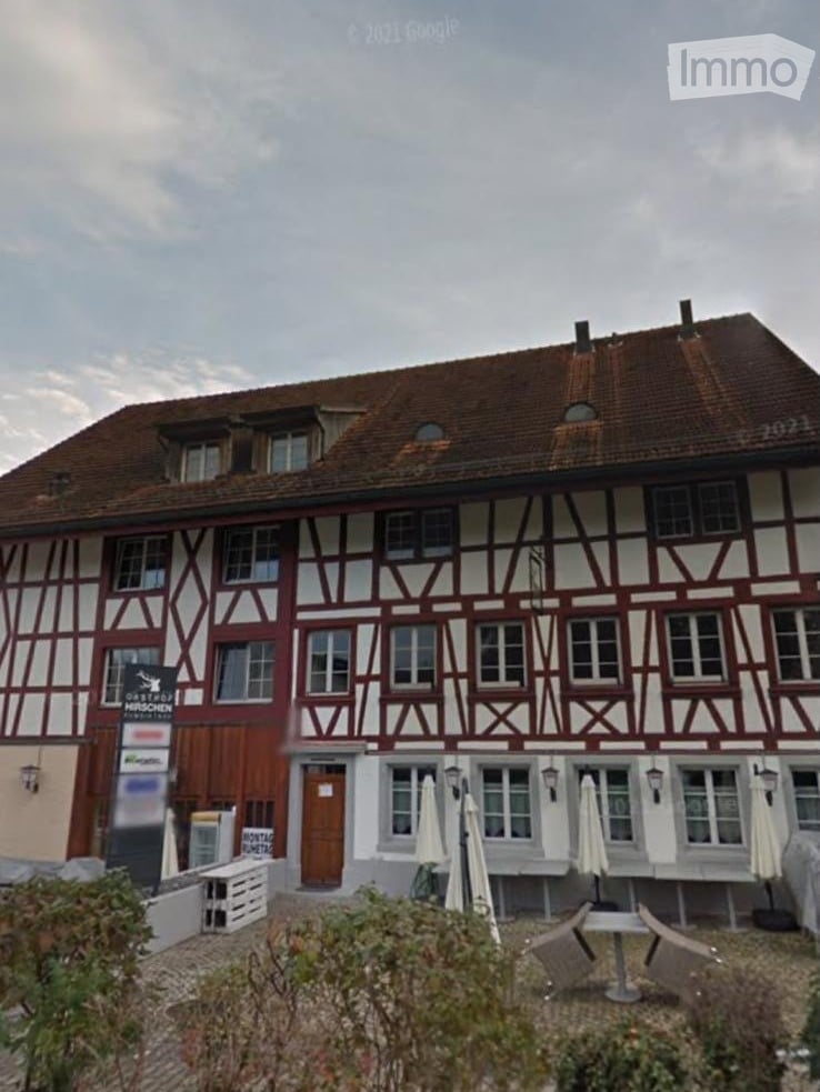 Traditional half-timbered building with red and white facade, multiple windows, and a brown tiled roof. Outdoor seating area with tables and chairs is visible in front.