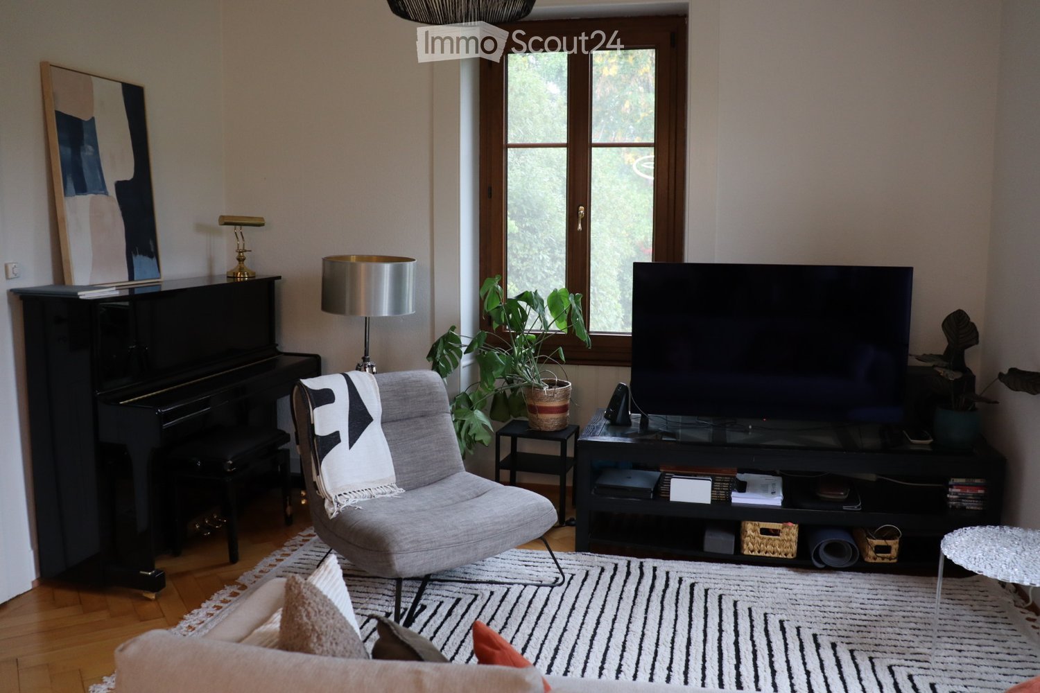 Modern living room with a grey sofa, a single armchair, a piano, a television, and a large window with a plant in front of it. The room features a patterned rug, a floor lamp, and a side table with a lamp.