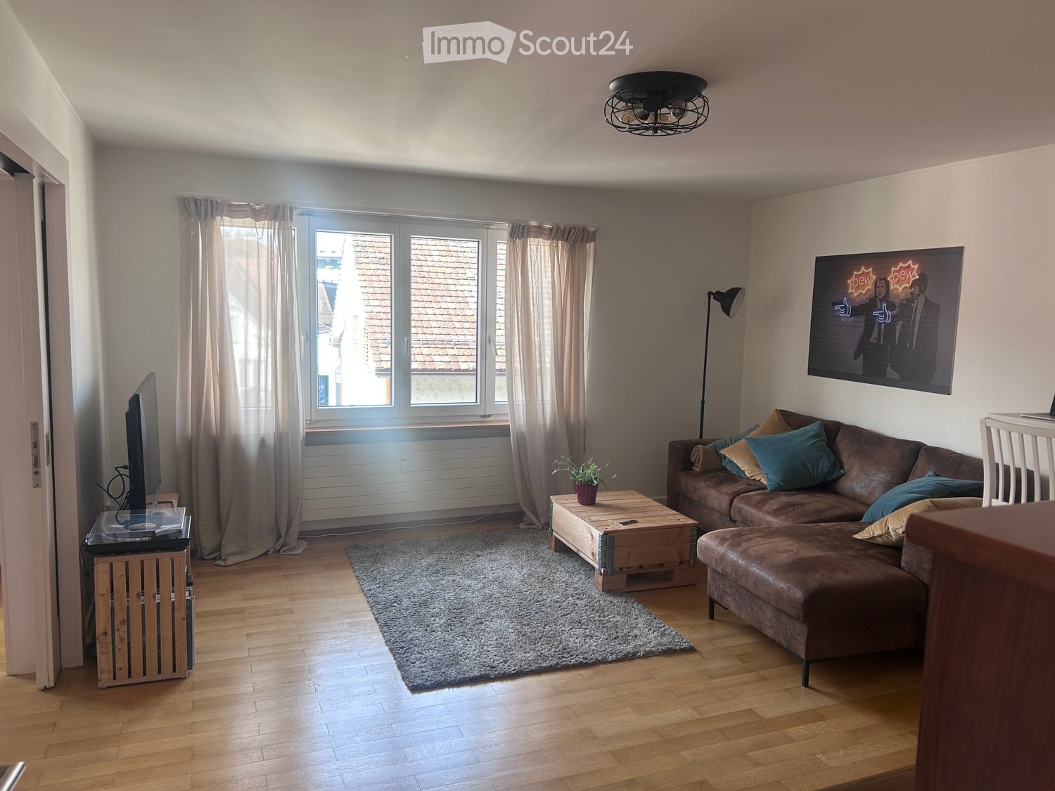 Modern living room with wooden flooring, large window with curtains, brown sectional sofa with multiple throw pillows, coffee table, TV on a stand, floor lamp, and a small plant on the coffee table.