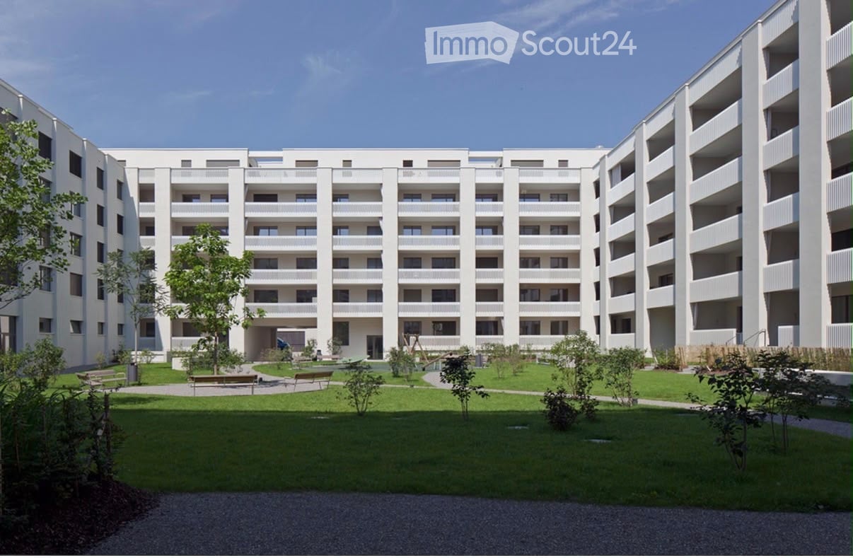 Modern multi-story residential building with white facade, multiple windows, and balconies. The courtyard features green lawns, trees, and benches.