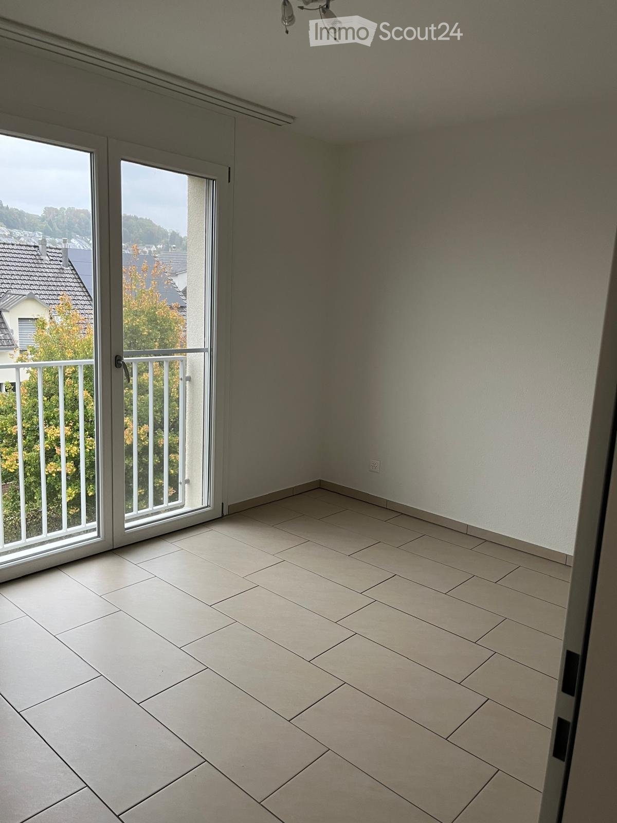Empty room with tiled floor, white walls, large sliding glass doors leading to a balcony with a metal railing and a view of the neighborhood.