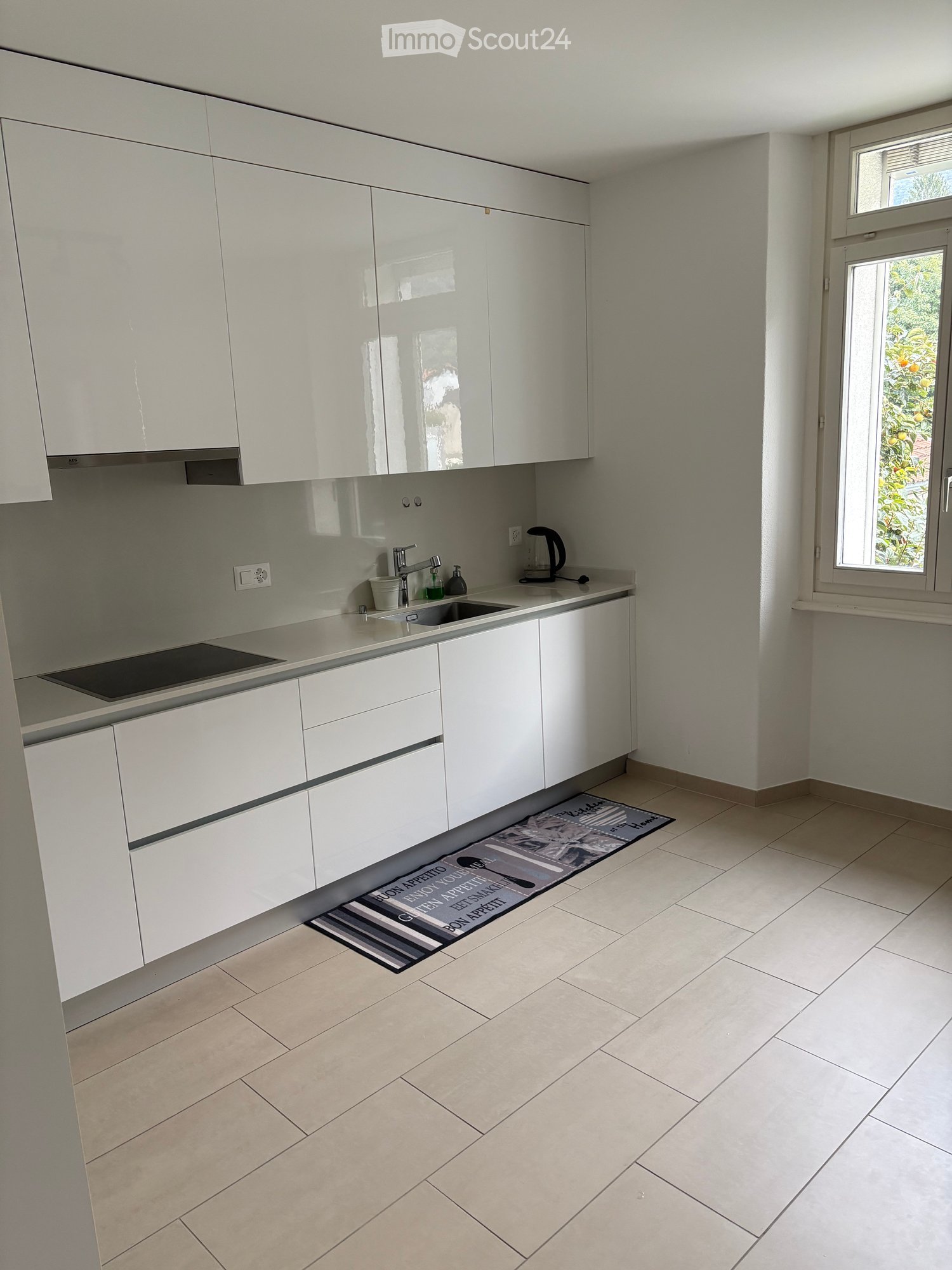 Modern kitchen with white cabinets, a sink, an induction cooktop, and a small rug on the floor. The room has tiled flooring and a window with a view of greenery.