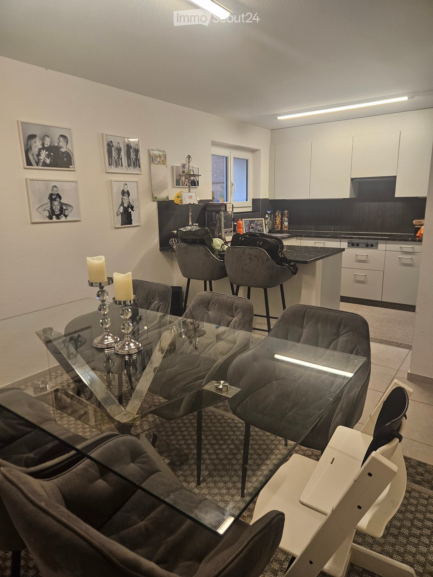 A modern, open-concept living and dining area with a glass dining table, grey upholstered chairs, and a small kitchen area with white cabinets and a black countertop.