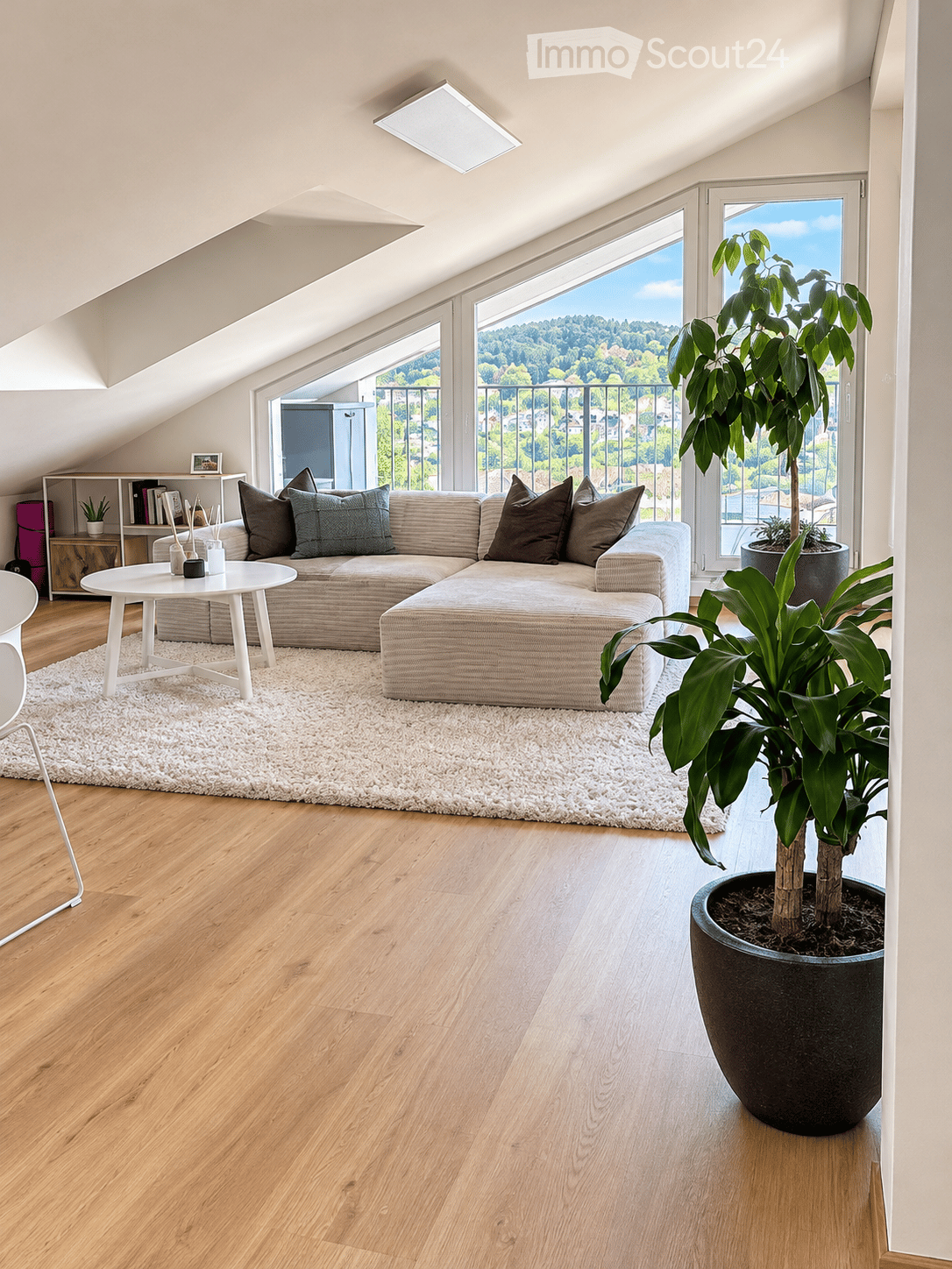 Modern living room with wooden flooring, large windows offering a scenic view, a sectional sofa, coffee table, and potted plants.