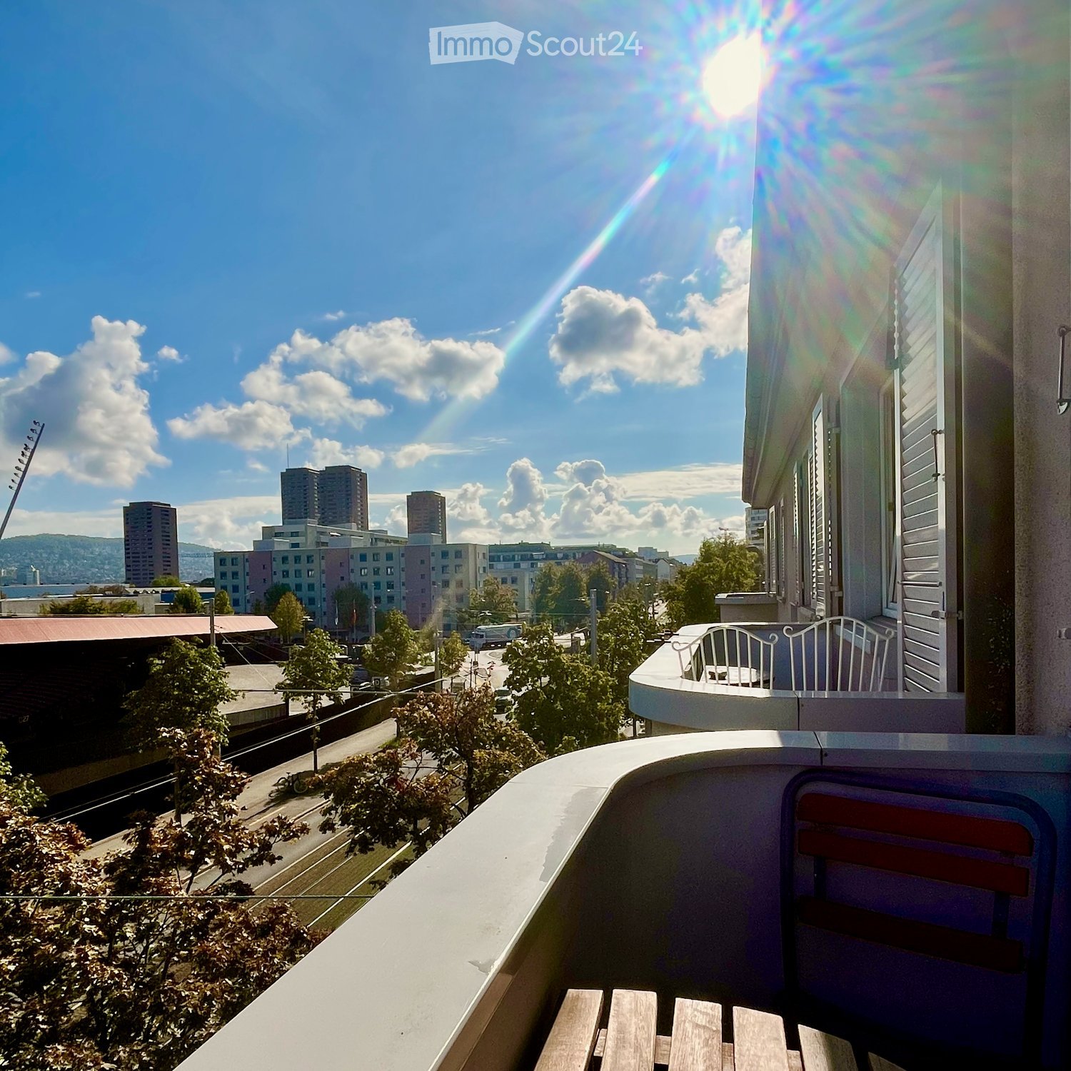 Balcony with wooden chairs and a table, overlooking a cityscape with buildings and trees.