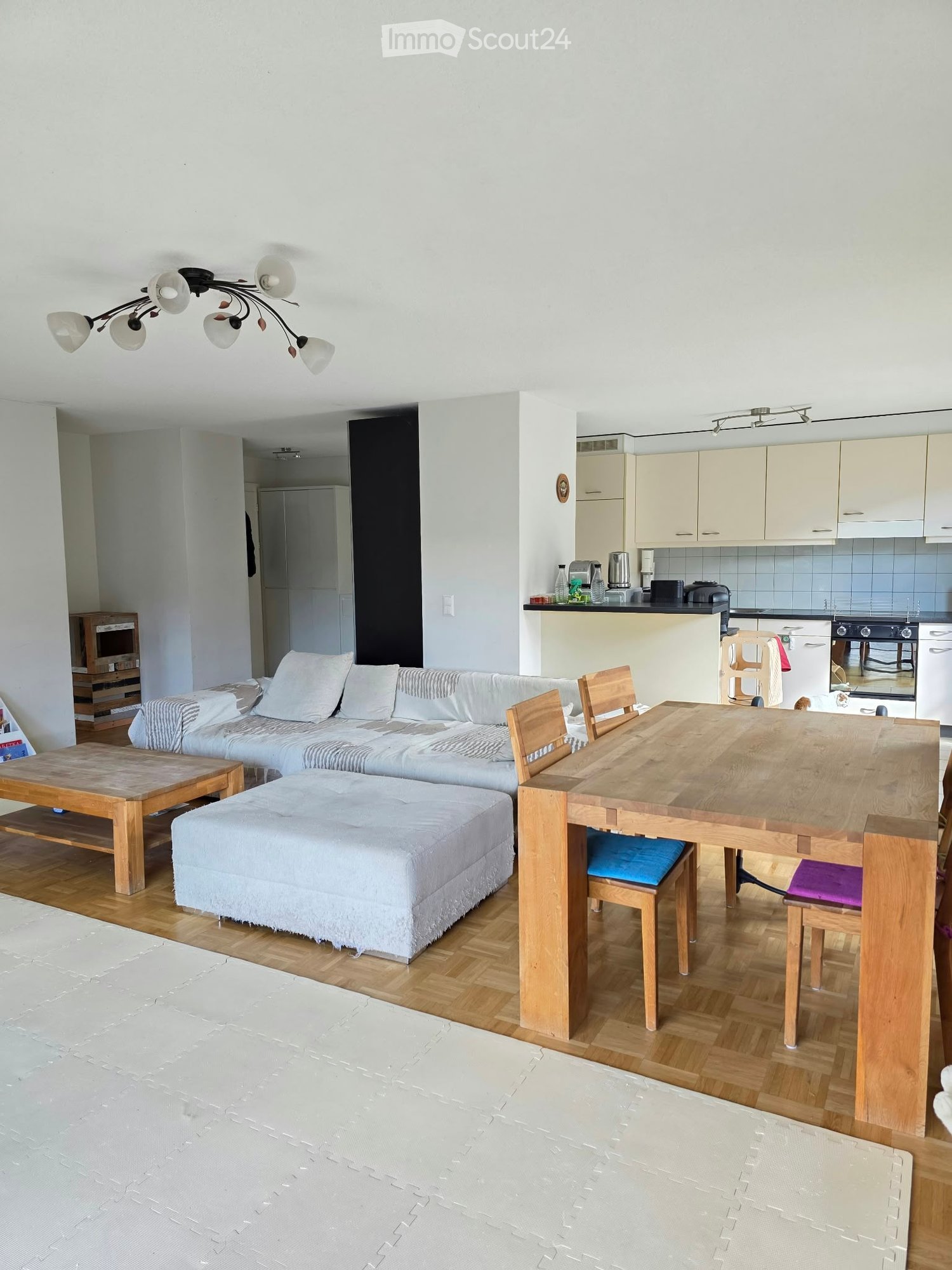 Bright, open-plan living and dining area with wooden flooring, white walls, and modern furniture including a sofa, ottoman, coffee table, dining table with chairs, and a small kitchen area with cabinets and appliances.