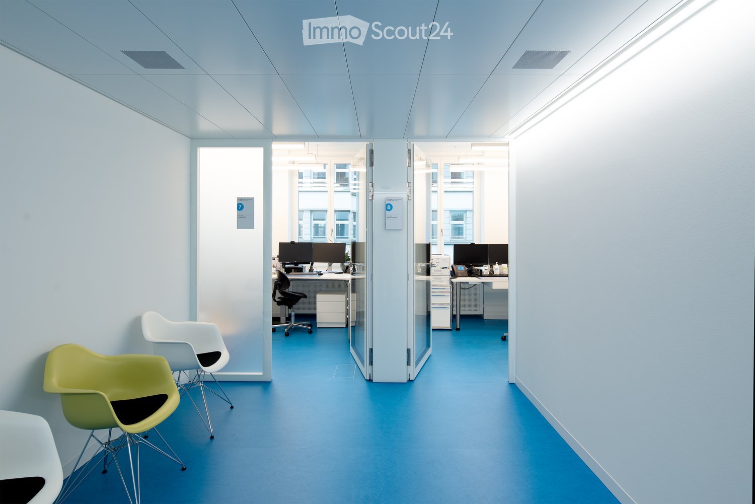 Modern office corridor with light blue walls and floor, featuring white and green chairs, desks with computers, and glass partitions.
