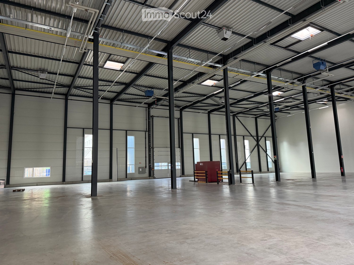 Large, empty industrial-style space with a high ceiling, metal beams, and large windows. The floor is concrete, and there are a few storage units and a small table in the center.
