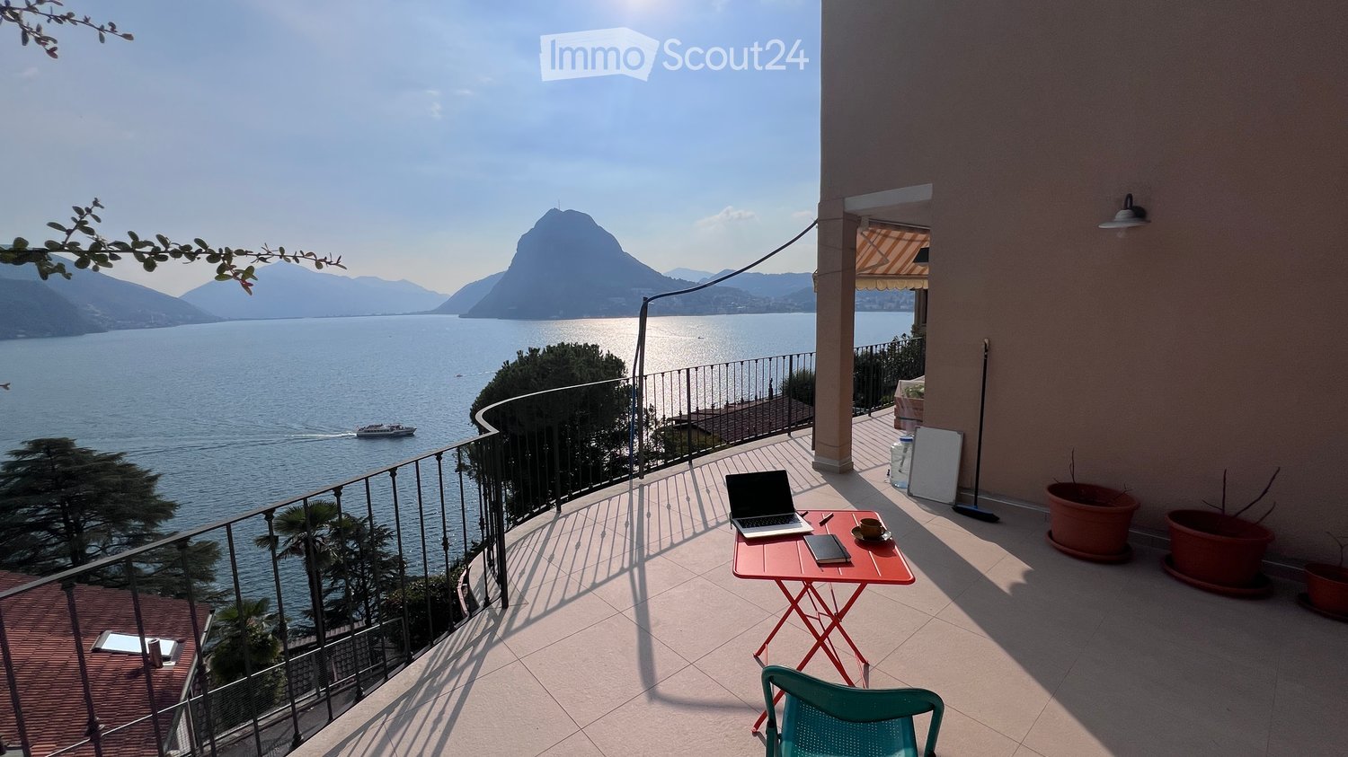 Balcony with a scenic lake view, featuring a small red table with a laptop, cup, and notebook, and a green plastic chair.
