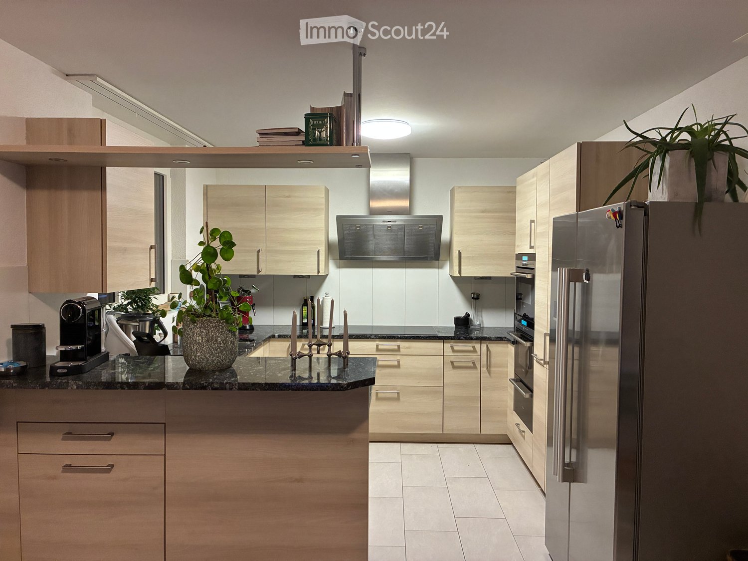 Modern kitchen with light wood cabinets, stainless steel appliances, granite countertop, and decorative items including a plant and candles.