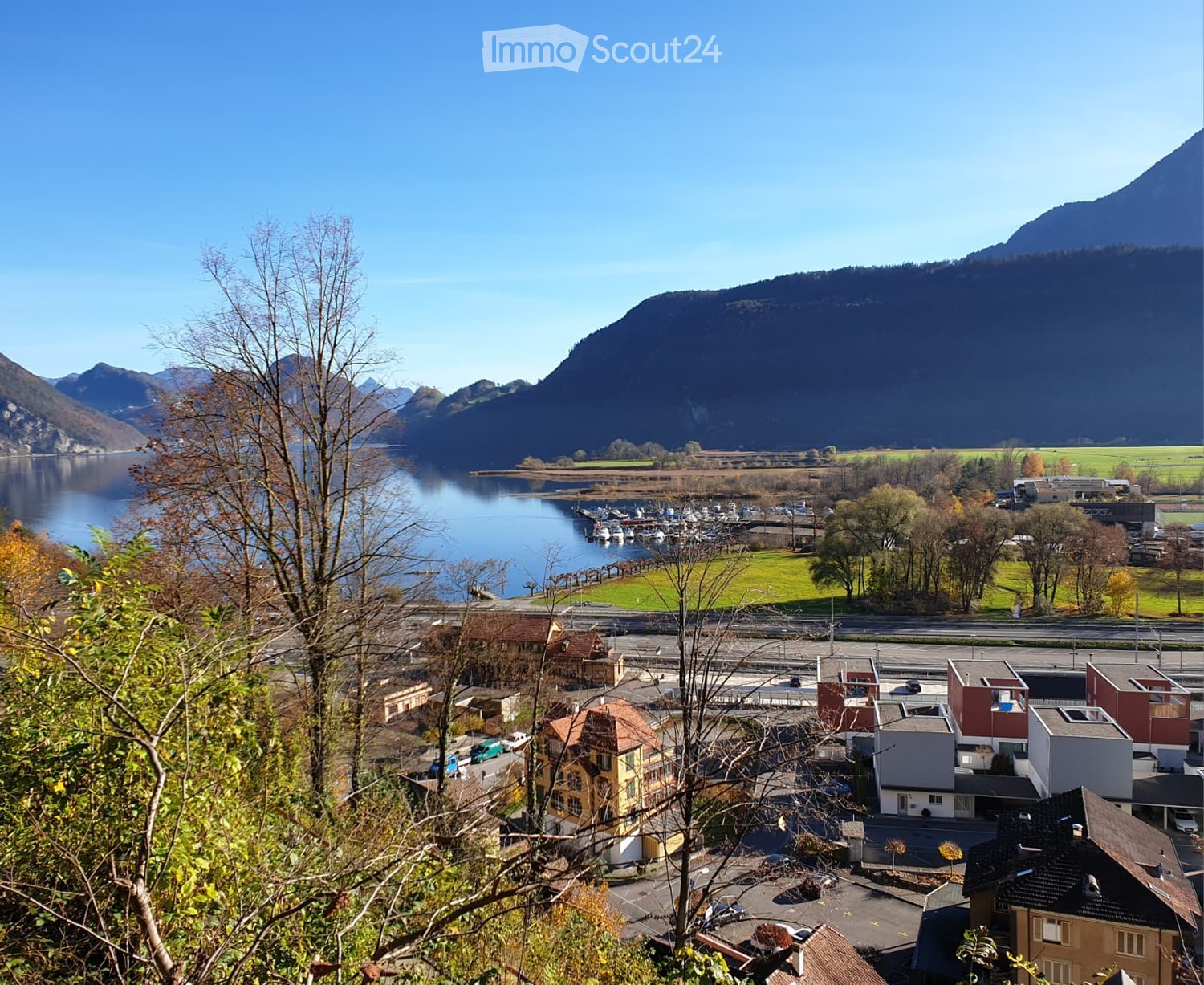 A scenic view of a town by a lake, surrounded by mountains, with houses, a marina, and a road visible.