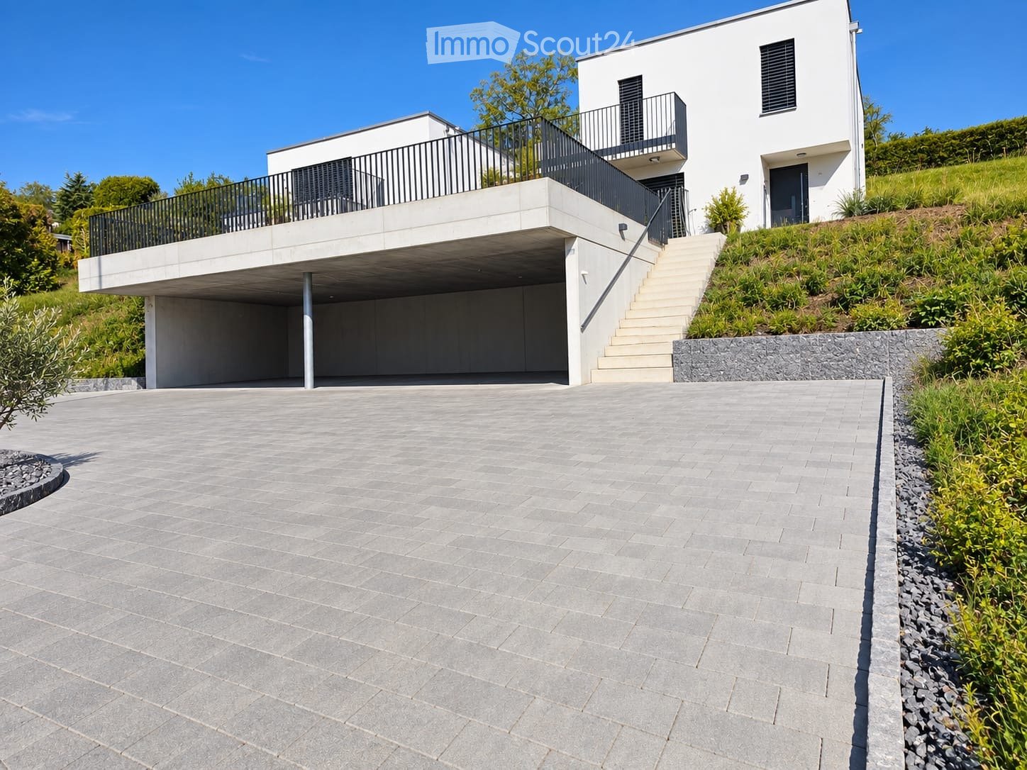 Modern two-story house with white exterior, raised ground floor, garage, balcony, and landscaped garden area.