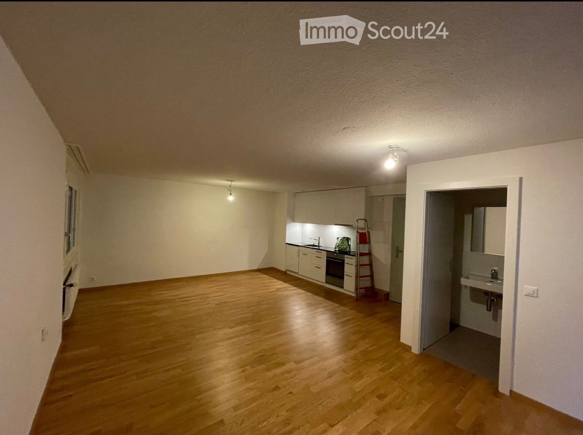 Empty room with wooden flooring, white walls, and a small kitchen area visible in the background. No furniture or personal items are present.