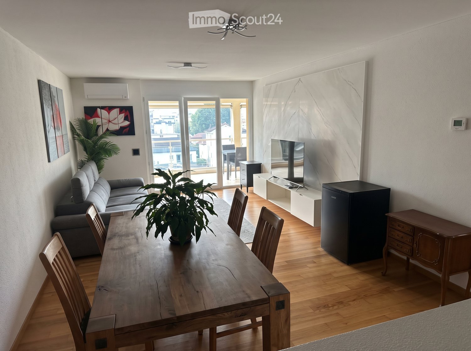 Modern living and dining area with a grey sofa, wooden dining table, chairs, a refrigerator, TV, and a balcony view.