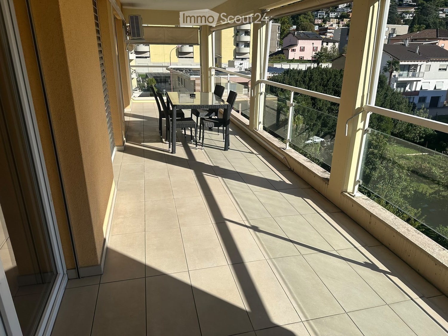Balcony with tiled floor, glass railing, and outdoor furniture including a table and chairs, overlooking a residential area with buildings and greenery.