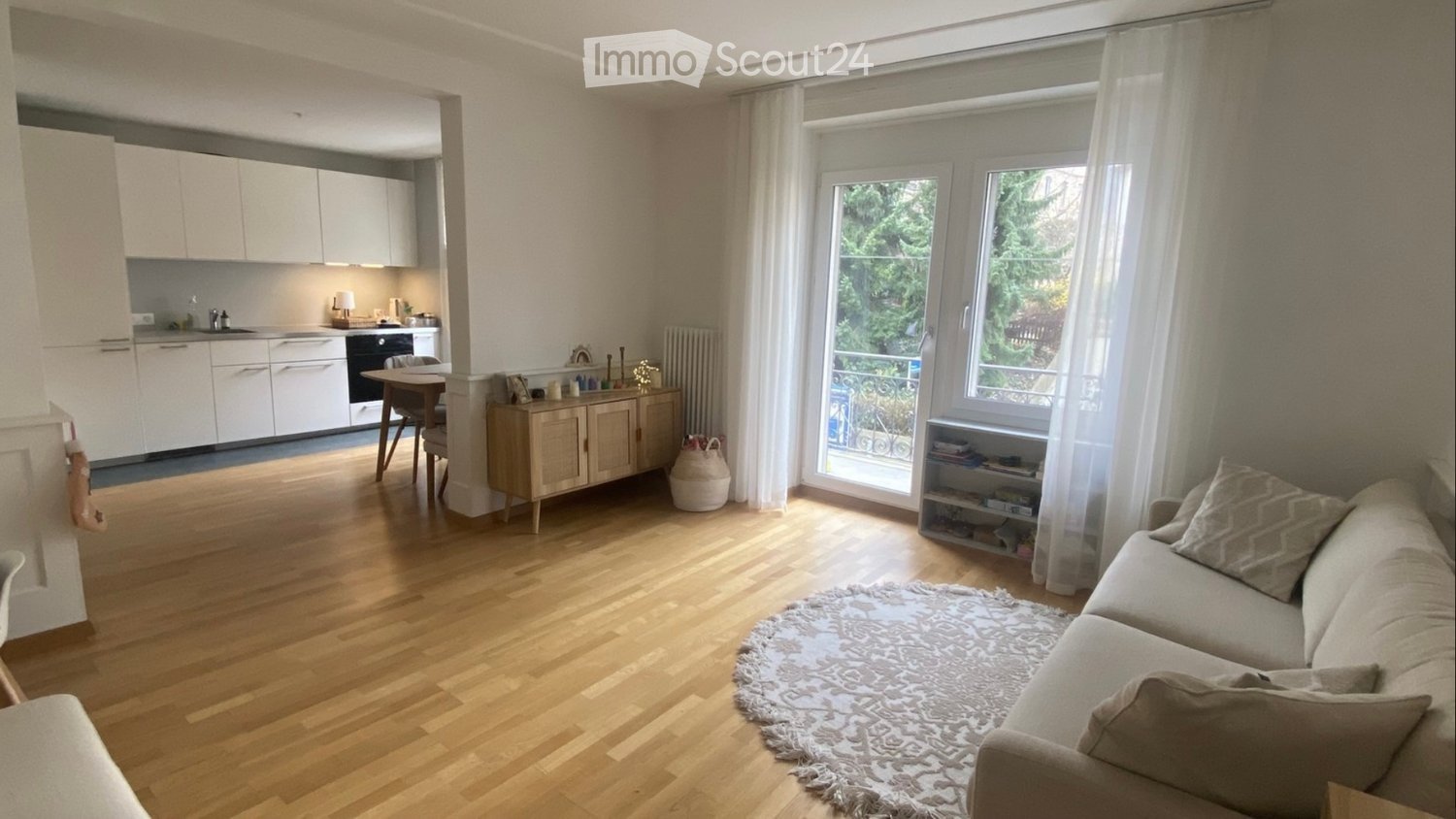 Bright living room with wooden flooring, large windows, a sofa, coffee table, and a small kitchen area with white cabinets and appliances.