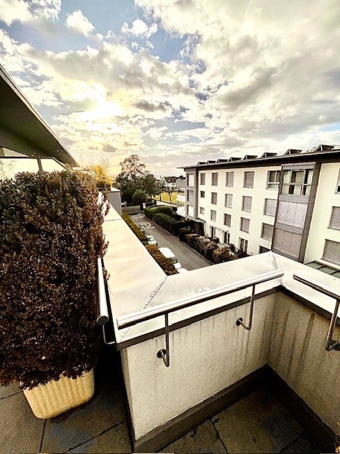 Balcony with metal railing, view of a multi-story building and parking area, greenery on the left side.