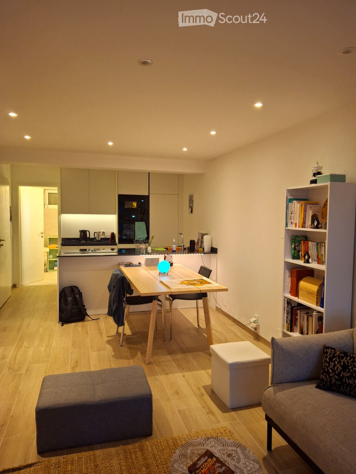 A modern, well-lit living space with a sofa, two stools, a small table with a lamp, a bookshelf filled with books, and a kitchen area with an oven and countertop.